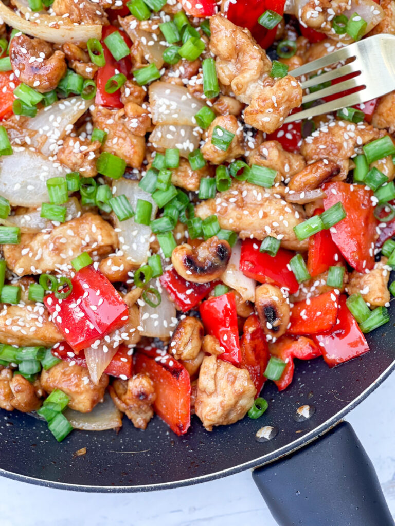 A photo of a stir-fry in a pan with veggies, chicken, and cashews. Topped with green onions.