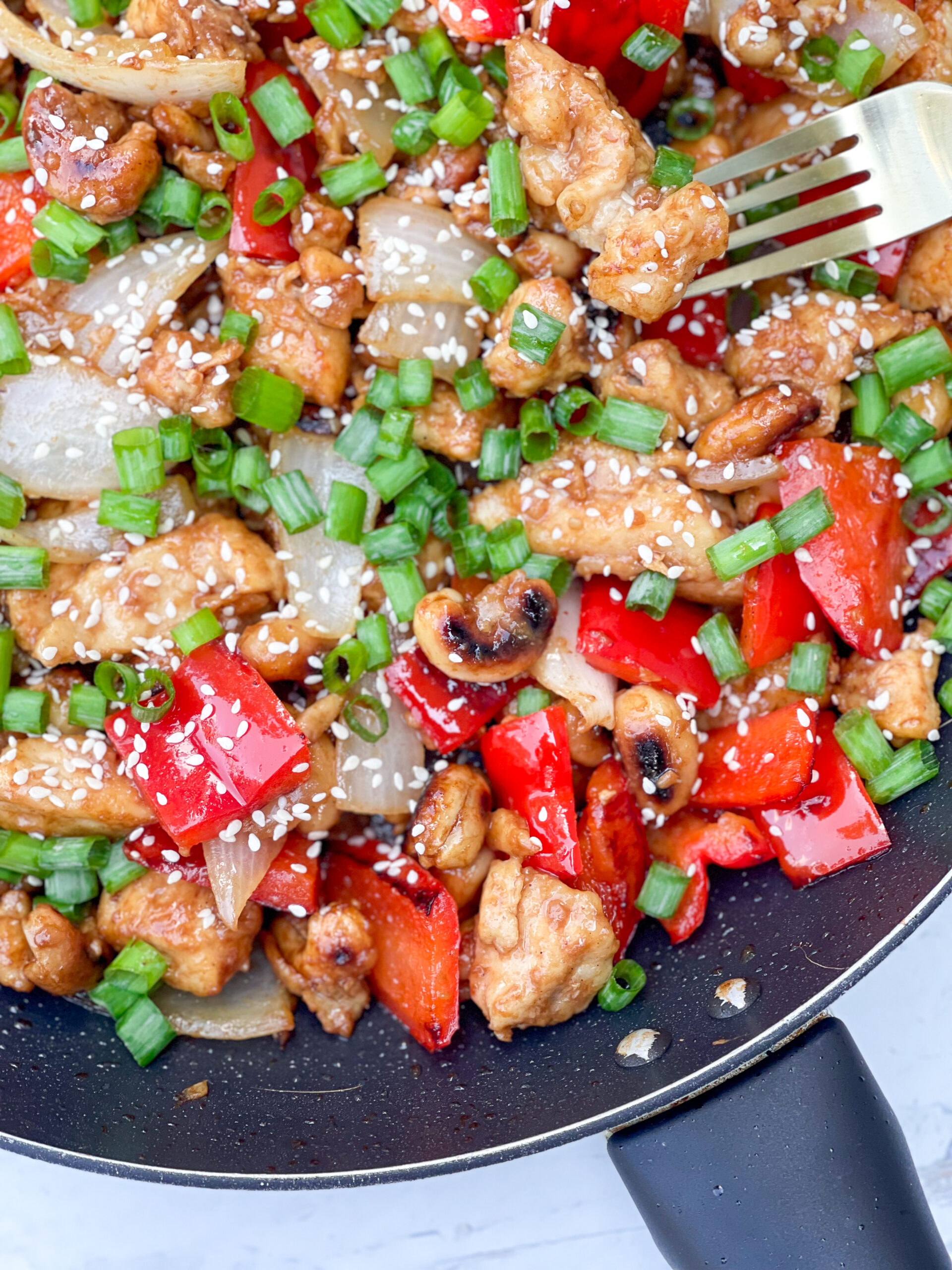 A photo of a stir-fry in a pan with veggies, chicken, and cashews. Topped with green onions.